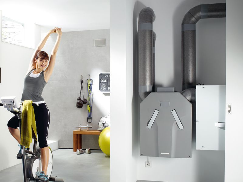 Frau macht Dehnübungen auf einem Heimtrainer im Fitnessraum, rechts moderne zentrale Wohnraumlüftungsanlage an der Wand.